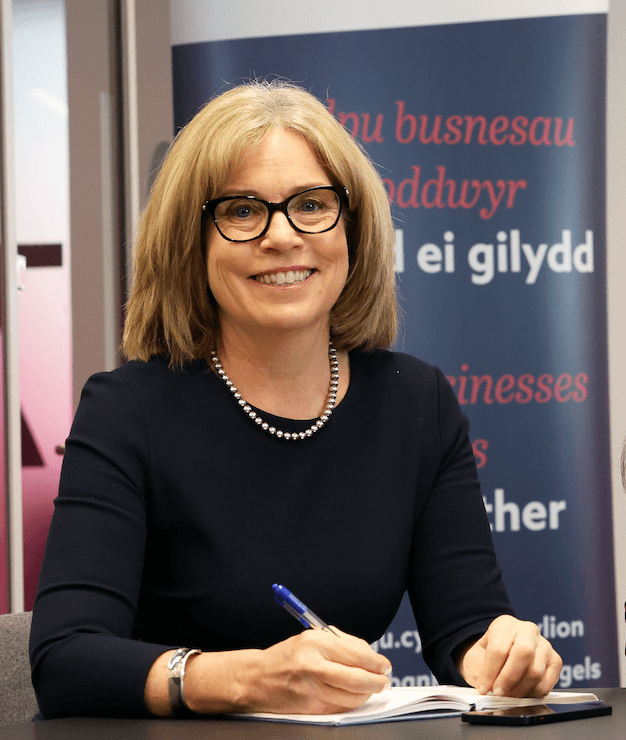 Smiling white woman with blonde hair. Wearing a black top, black framed glasses and pearls while writing in a notebook.