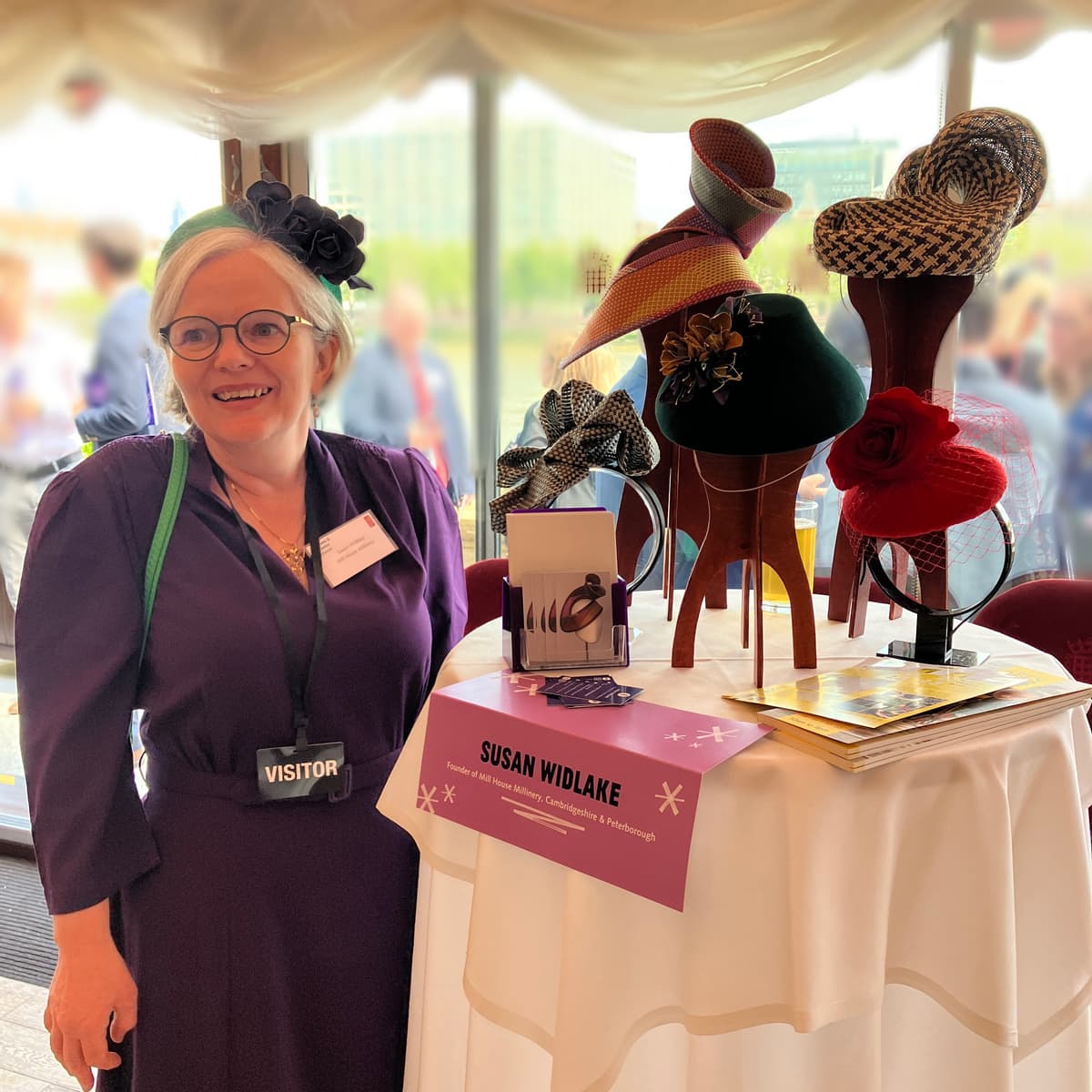 Woman in purple top. Standing beside headwear and hats on a table.